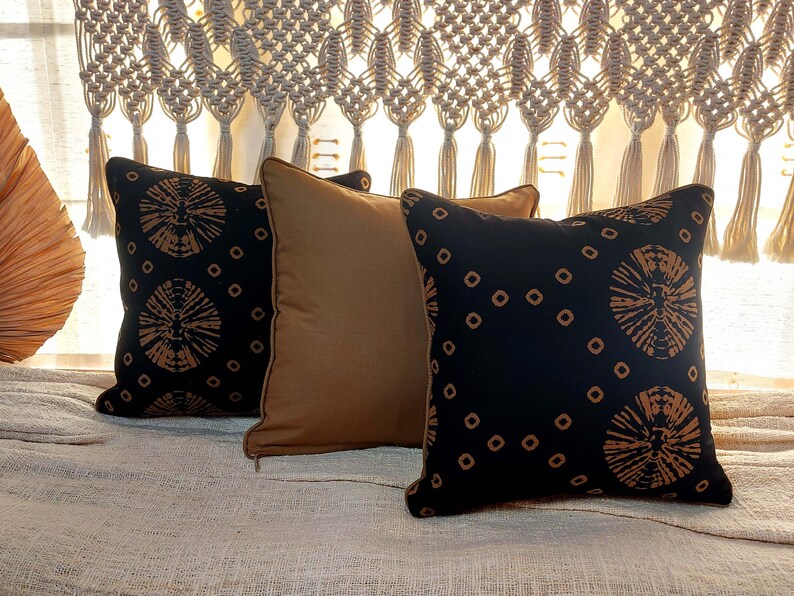 May include: Three decorative throw pillows with a black background and a brown geometric pattern. The pillows are arranged on a beige textured surface.