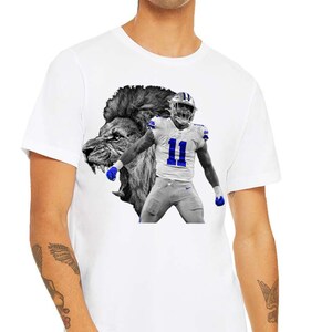 May include: White t-shirt featuring a graphic of a football player in a blue and white uniform with the number 11, superimposed over a black and white lion's head. The shirt is a crew neck style.