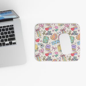 May include: A rectangular mouse pad featuring a repeating pattern of pastel-colored stationery items including pencils, scissors, notebooks, paint palettes, and rainbows, with the word hello written in cursive script, topped with a white computer mouse.