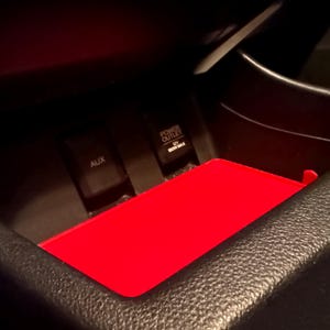 May include: A bright red rectangular tray insert positioned within a black vehicle center console storage compartment. The compartment features an AUX port and a 12V power outlet labeled 120W MAX. The tray provides a flat surface for storage.