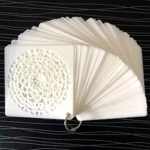 Op de afbeelding: Een witte plastic sjabloon met een cirkelvormig ontwerp. De sjabloon bestaat uit meerdere kaarten die met een metalen ring aan elkaar zijn verbonden.