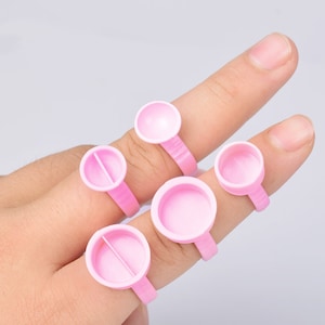 Puede incluir: Cinco anillos de dedo rosas con pequeños recipientes redondos en la parte superior. Los anillos están diseñados para contener pequeñas cantidades de líquido, como pegamento o esmalte de uñas.