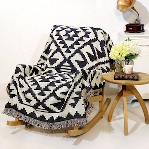 May include: A black and white geometric patterned throw blanket draped over a rocking chair. The blanket has fringe on the edges. The chair is made of wood and has a natural finish.