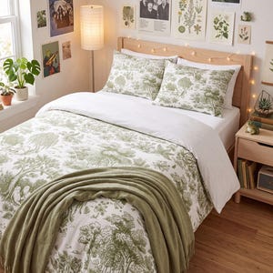May include: A bedding set featuring a white duvet cover and two matching pillowcases with a green toile-style botanical print of trees and foliage. A solid olive green throw blanket is draped across the foot of the bed.