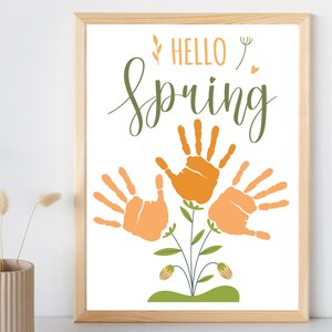 May include: A framed art print with a light wood frame. The print features the words "HELLO SPRING" in green script above a floral design made of orange handprints. The handprints are arranged to resemble flowers.