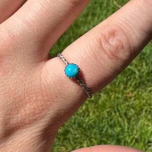 May include: A silver ring with a twisted band and a round, bright turquoise stone. The ring is set against a background of green grass.