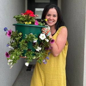 May include: A woman in a yellow jumpsuit holds a green hanging planter filled with colorful flowers. The planter has multiple holes for planting. The flowers are pink, purple, white, and red.
