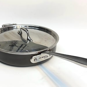 May include: A dark gray All-Clad frying pan with a stainless steel handle and lid. The pan has a clear glass lid with a stainless steel handle. The handle is long and curved. The pan is on a white background.