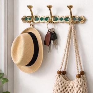 Puede incluir: Un perchero de pared de latón con tres ganchos curvos. El perchero presenta un patrón de diamantes verdes y blancos. Un sombrero de paja con una banda negra, llaves y una bolsa de macramé beige cuelgan de los ganchos.