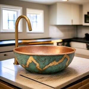 May include: A copper sink with a teal and gold finish sits on a countertop, paired with a gold faucet. The sink's interior is a warm copper color, and the exterior features a unique teal patina with gold accents. The faucet has a curved design.
