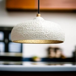May include: A white, textured pendant light with a gold-coloured interior. The light fixture is suspended by a black cord and has a gold-coloured accent at the top. The light has a unique, dimpled surface.
