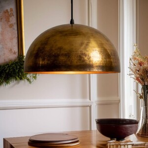 Peut inclure: Un grand luminaire suspendu en forme de dôme doré est suspendu à un cordon noir. Le luminaire est suspendu au-dessus d'une table en bois, avec un bol foncé et des objets décoratifs visibles en dessous. L'intérieur de la lampe brille d'une lumière chaude.