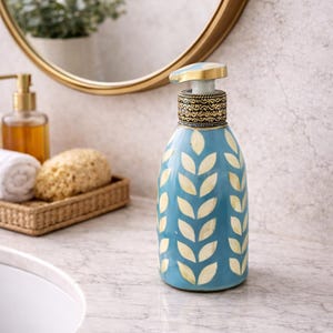 Peut inclure: Un distributeur de savon en céramique bleu avec un motif de feuilles crème. Le distributeur a une pompe dorée et une bande décorative. Il est posé sur un comptoir en marbre, avec un miroir et d'autres accessoires de salle de bain en arrière-plan.