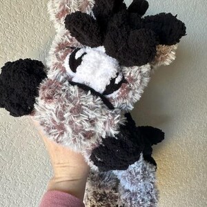 May include: A handmade plush toy in the shape of a cow. The toy features a fluffy, textured body with a mix of brown, white, and black yarn. The cow has black ears, eyes, and hooves, and a white face.