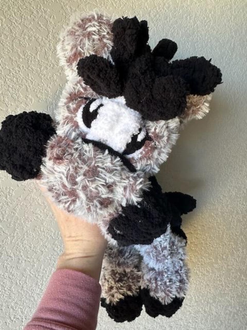 May include: A handmade plush toy in the shape of a cow. The toy features a fluffy, textured body with a mix of brown, white, and black yarn. The cow has black ears, eyes, and hooves, and a white face.
