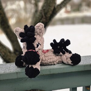 May include: A handmade, plush horse toy in tan and black. The horse has black hooves, a black mane and tail, and a pink mouth. The toy is lying on a green wooden railing covered in snow, with a blurred winter background.