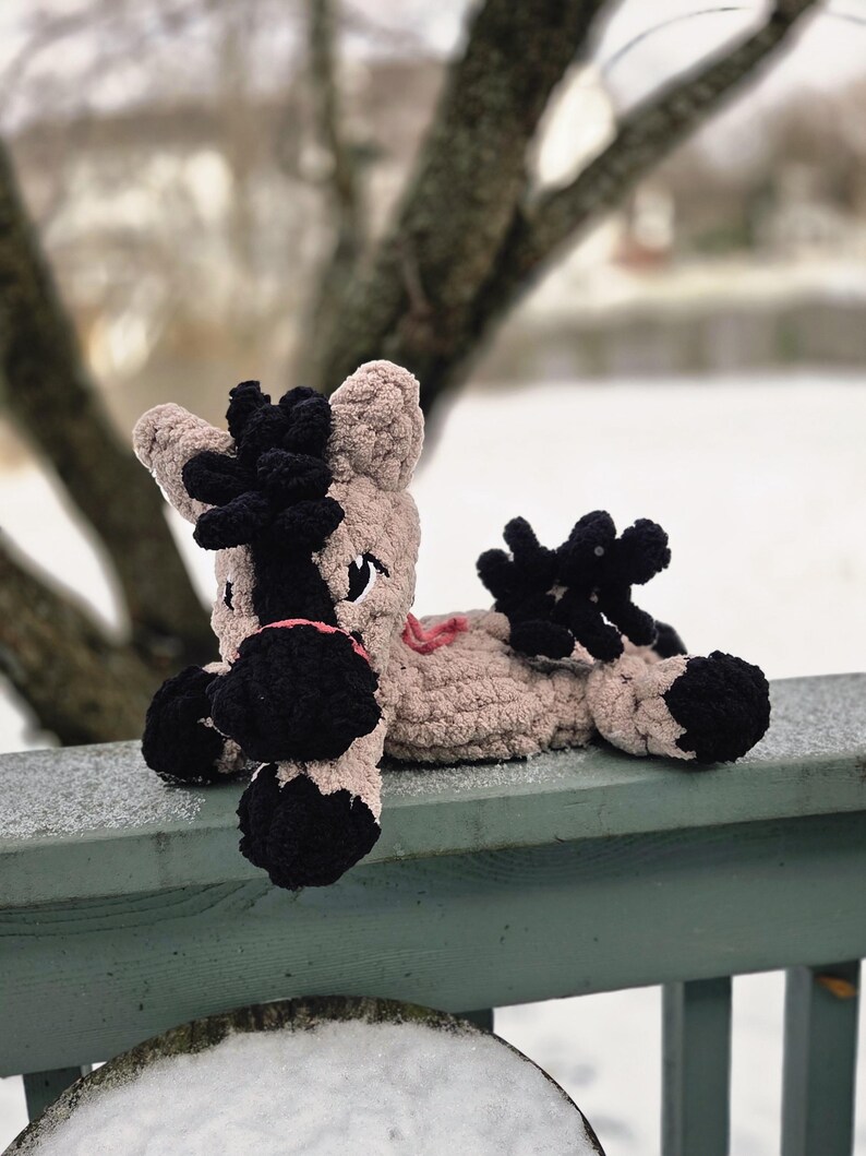 May include: A handmade, plush horse toy in tan and black. The horse has black hooves, a black mane and tail, and a pink mouth. The toy is lying on a green wooden railing covered in snow, with a blurred winter background.