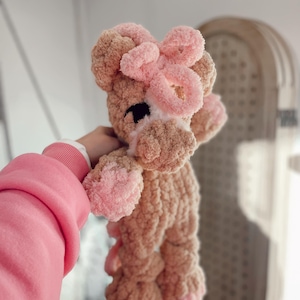 May include: A handmade, plush giraffe toy in shades of tan and pink. The giraffe has a pink bow on its head, pink hooves, and a white muzzle. The toy is made of a soft, textured material, perfect for a child's comfort.