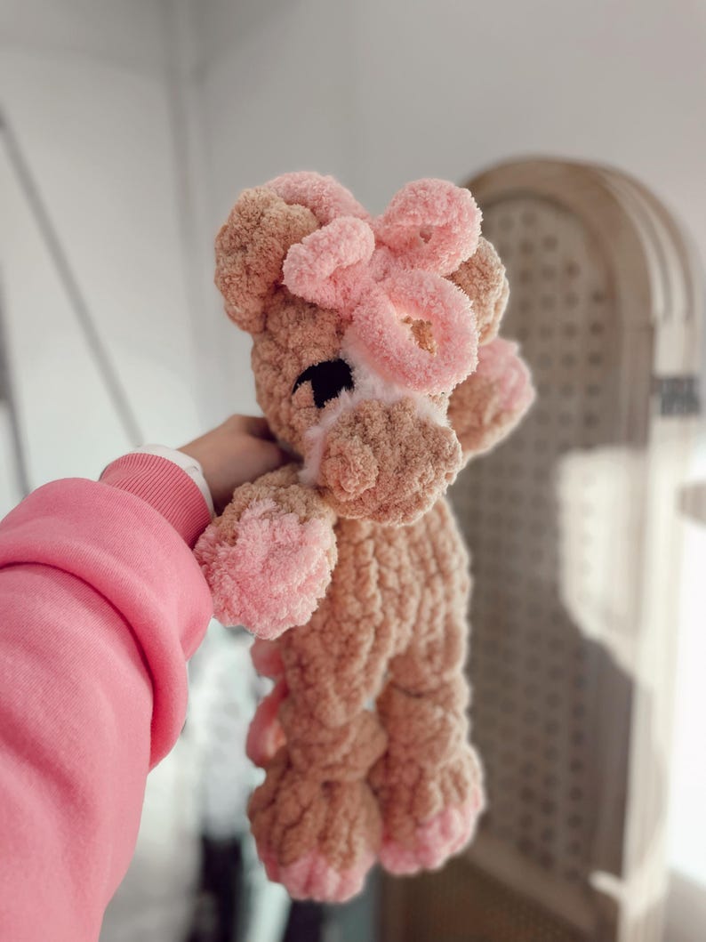 May include: A handmade, plush giraffe toy in shades of tan and pink. The giraffe has a pink bow on its head, pink hooves, and a white muzzle. The toy is made of a soft, textured material, perfect for a child's comfort.