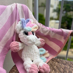 May include: A white crochet unicorn with pink and pastel accents, including a rainbow mane and hooves. The stuffed animal is seated on a pink and white striped towel, creating a whimsical scene. The unicorn is a handmade item.