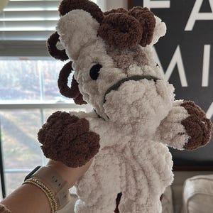 May include: A handmade plush cow toy with a fluffy white body and brown accents on the ears, hooves, and head. The cow has black eyes and a green halter. The toy is soft and cuddly, perfect for children.
