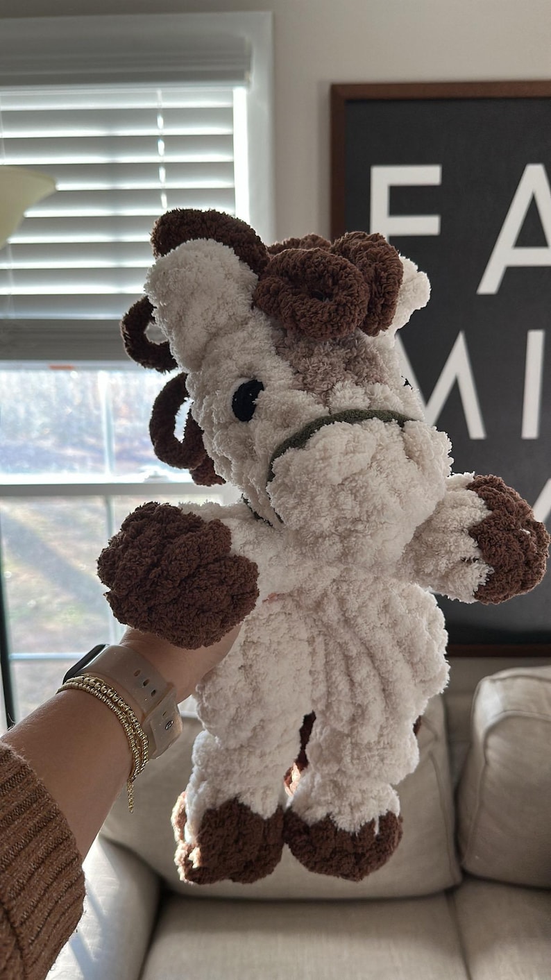 May include: A handmade plush cow toy with a fluffy white body and brown accents on the ears, hooves, and head. The cow has black eyes and a green halter. The toy is soft and cuddly, perfect for children.