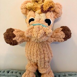 May include: A handmade plush giraffe toy in shades of tan, brown, and cream. The giraffe has large black eyes, a light blue mouth, and a fluffy mane. The toy is made of soft, textured yarn and has outstretched arms and legs.