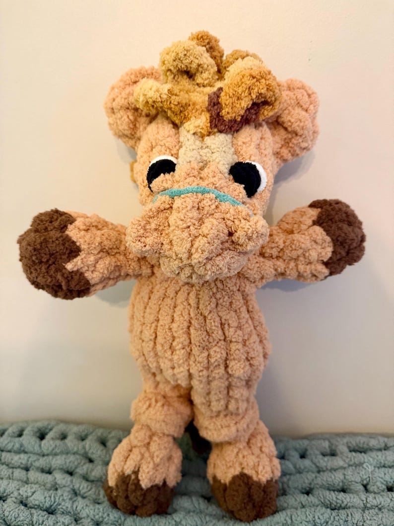 May include: A handmade plush giraffe toy in shades of tan, brown, and cream. The giraffe has large black eyes, a light blue mouth, and a fluffy mane. The toy is made of soft, textured yarn and has outstretched arms and legs.