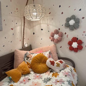 May include: A white bed with a floral patterned duvet cover and a variety of pillows, including a round yellow pillow, a heart-shaped pillow, and a velvet pillow. The bed is in a room with white walls and a white ceiling. There are three crocheted flower decorations on the wall.
