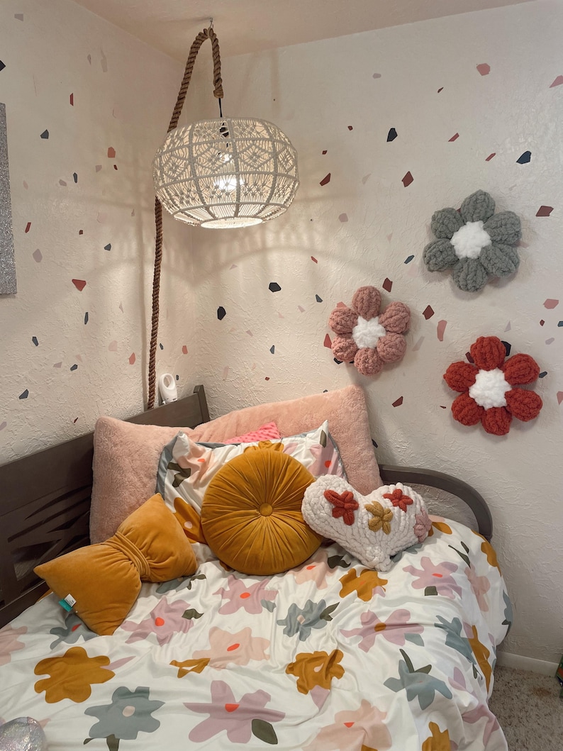 May include: A white bed with a floral patterned duvet cover and a variety of pillows, including a round yellow pillow, a heart-shaped pillow, and a velvet pillow. The bed is in a room with white walls and a white ceiling. There are three crocheted flower decorations on the wall.