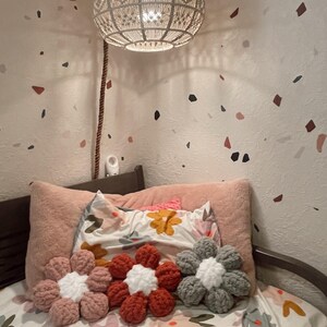May include: Three crocheted flower-shaped pillows in pink, red, and gray on a bed with a floral patterned duvet cover. The pillows are arranged in a row on the bed.