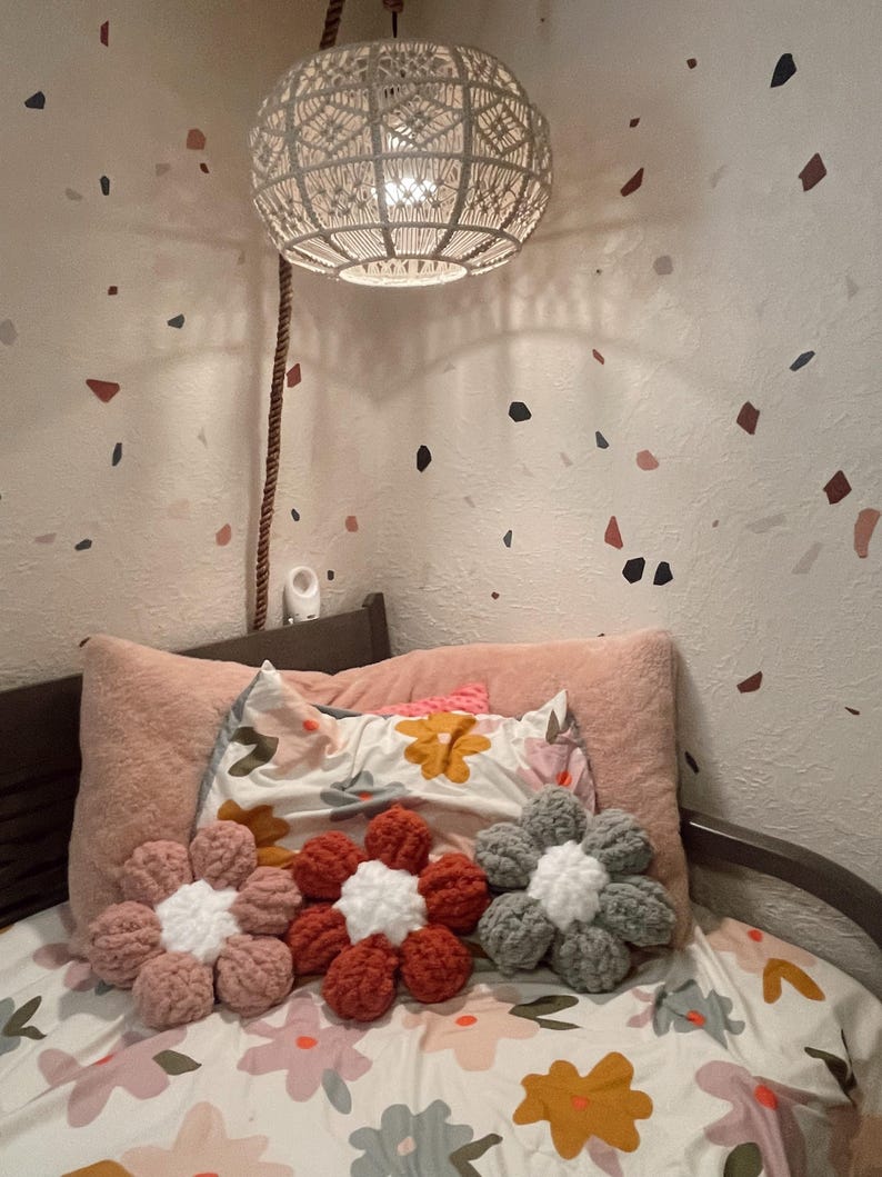 May include: Three crocheted flower-shaped pillows in pink, red, and gray on a bed with a floral patterned duvet cover. The pillows are arranged in a row on the bed.