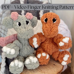 May include: Two plush finger-knitted dinosaur toys. One is gray with pink accents and white paws, the other is orange with white paw pads. The toys are sitting on a wooden surface.