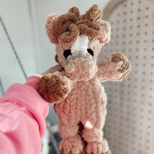 May include: A handmade plush horse toy in shades of brown and tan. The horse has a white patch on its face, black eyes, and a light blue mouth. The toy is made of soft, textured yarn and has a fluffy mane and tail.