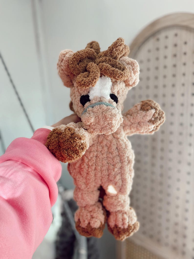 May include: A handmade plush horse toy in shades of brown and tan. The horse has a white patch on its face, black eyes, and a light blue mouth. The toy is made of soft, textured yarn and has a fluffy mane and tail.