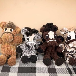 May include: Four handmade crochet horse plush toys in various colors. The horses have detailed features, including black eyes, manes, and hooves. The color variations include brown, gray, and a mix of brown and white. They are arranged on a checkered surface.