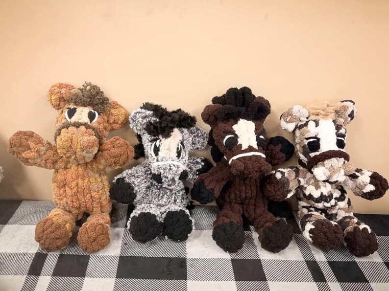 May include: Four handmade crochet horse plush toys in various colors. The horses have detailed features, including black eyes, manes, and hooves. The color variations include brown, gray, and a mix of brown and white. They are arranged on a checkered surface.