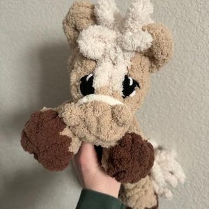 May include: A handmade plush horse toy in shades of tan, brown, and cream. The horse has large black eyes, a white mane, and brown hooves. The soft, cuddly toy is held in a person's hand.