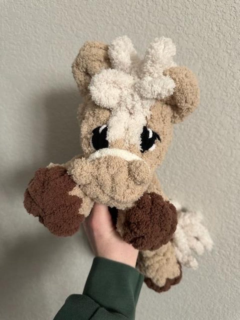 May include: A handmade plush horse toy in shades of tan, brown, and cream. The horse has large black eyes, a white mane, and brown hooves. The soft, cuddly toy is held in a person's hand.