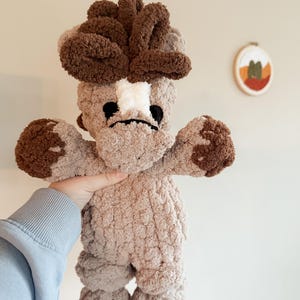May include: A handmade plush toy in the shape of a mushroom. The mushroom has a light brown body with darker brown accents on the head, arms, and feet. The face features black eyes and a white patch. The toy is held up by a person.