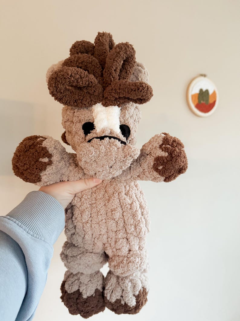 May include: A handmade plush toy in the shape of a mushroom. The mushroom has a light brown body with darker brown accents on the head, arms, and feet. The face features black eyes and a white patch. The toy is held up by a person.