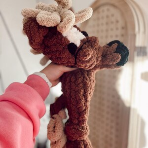 May include: A handmade brown and beige crochet deer plushie with a white patch on its face and black hooves. The deer has large, fluffy antlers and is made with a soft, textured yarn. A unique, handcrafted item.
