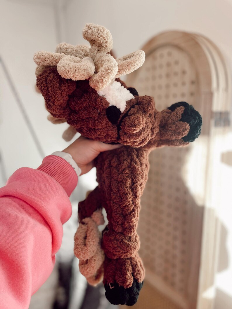 May include: A handmade brown and beige crochet deer plushie with a white patch on its face and black hooves. The deer has large, fluffy antlers and is made with a soft, textured yarn. A unique, handcrafted item.