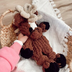 May include: A handmade brown and tan plush reindeer toy with black hooves and antlers. The reindeer is lying in a woven basket on a white blanket. The plush toy is made of soft, textured yarn.