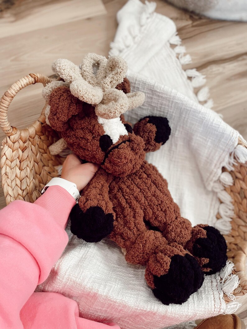 May include: A handmade brown and tan plush reindeer toy with black hooves and antlers. The reindeer is lying in a woven basket on a white blanket. The plush toy is made of soft, textured yarn.