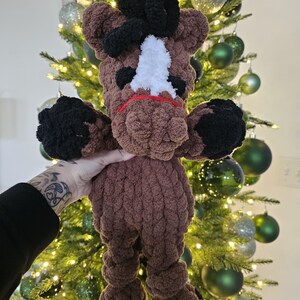 May include: A handmade brown and black crochet horse plushie with a white face marking and red accents. The plushie has black hooves and a black mane. The horse is being held in front of a Christmas tree with green and silver ornaments.