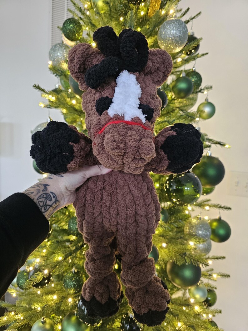 May include: A handmade brown and black crochet horse plushie with a white face marking and red accents. The plushie has black hooves and a black mane. The horse is being held in front of a Christmas tree with green and silver ornaments.
