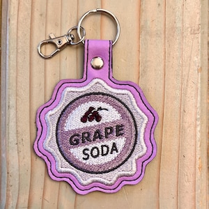 May include: Purple leather keychain with a white and purple embroidered grape soda bottle cap design. The keychain has a silver metal ring and a silver metal clasp.