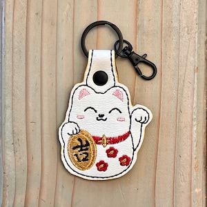 May include: White lucky cat keychain with a red collar and a gold coin with the Japanese character for "fortune" on it. The cat is holding its right paw up in the air.