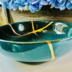 Puede incluir: Un cuenco de cerámica azul verdoso con líneas de craquelado doradas. El cuenco es redondo y tiene un acabado liso y brillante.
